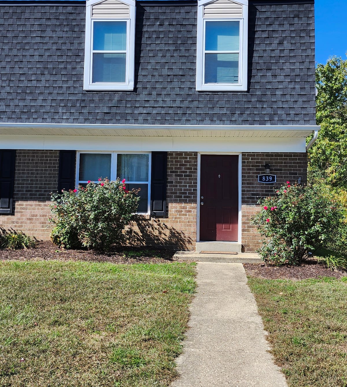 The brick front of a Calvertowne townhouse under a clear blue sky, with a maroon front door, rose bushes, and a concrete walkway across the lawn
