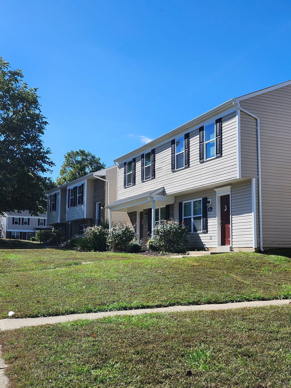 A row of two-story townhouses at Calvertowne with tan siding, dark shutters, and a mown lawn beside the sidewalk