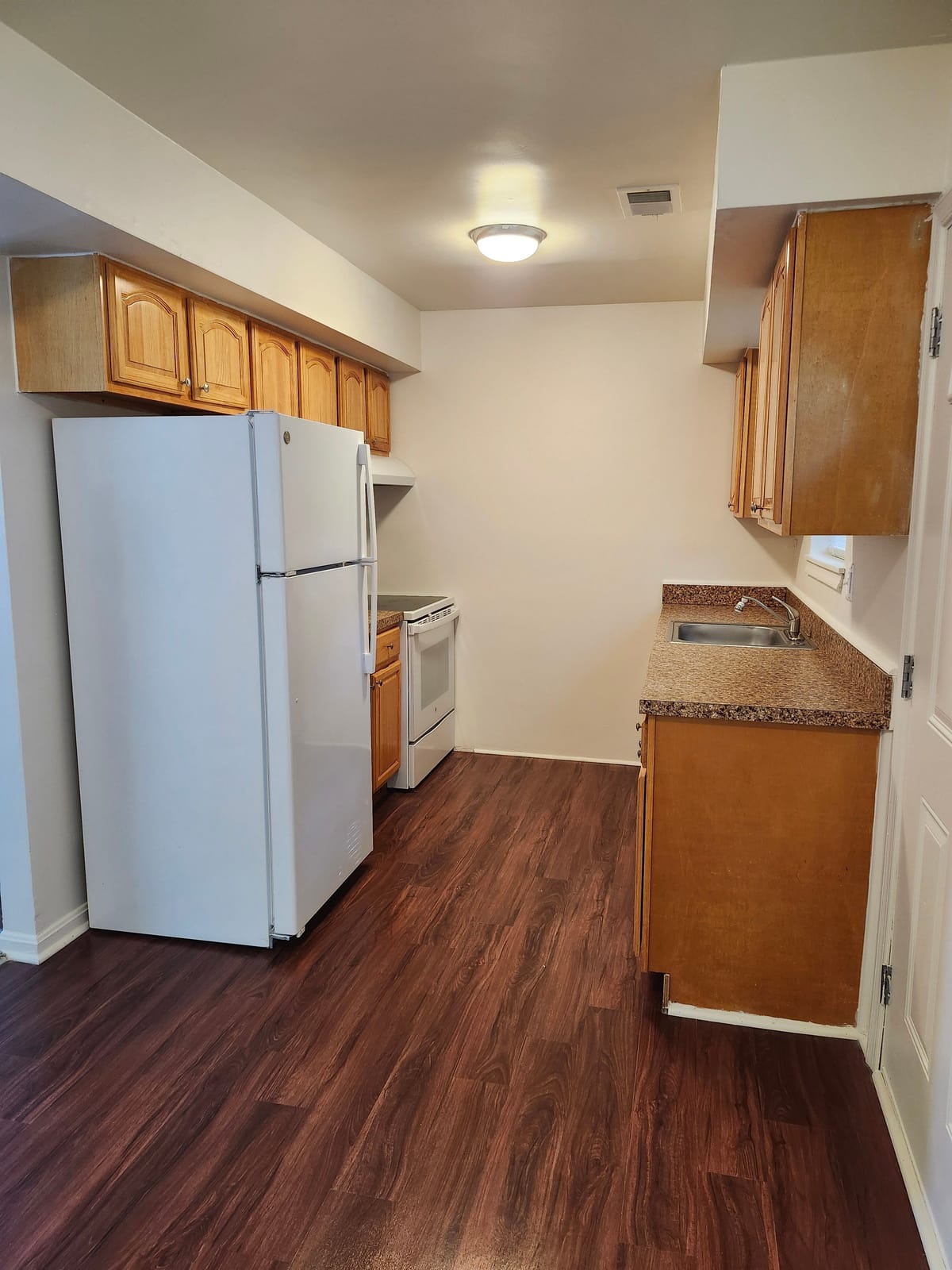 A galley kitchen at Calvertowne with oak cabinets, a white refrigerator and electric range, a double stainless sink, and dark wood-look plank flooring
