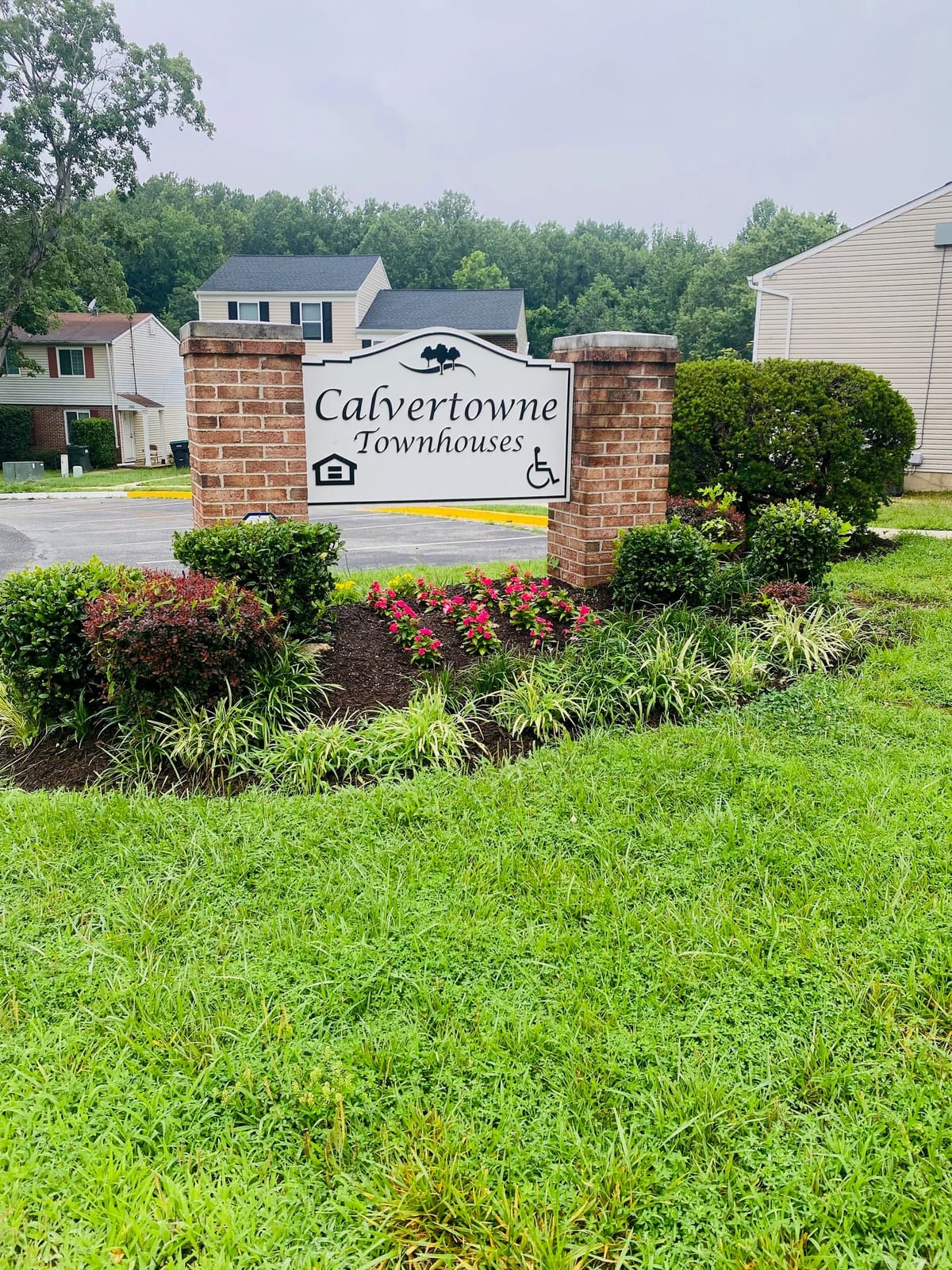 The Calvertowne Townhouses entrance sign on brick piers, above a mulched bed of pink flowers, with townhouses and trees behind it
